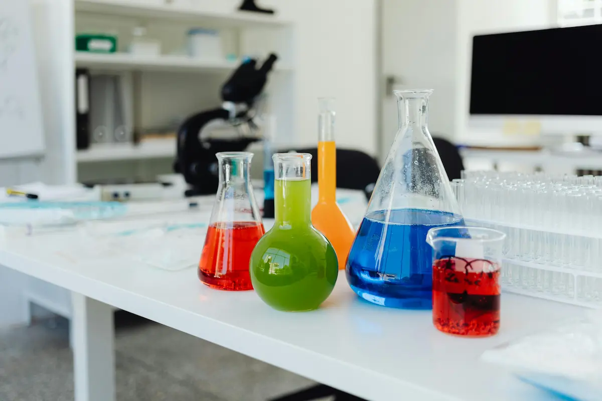 Colorful chemistry flasks filled with red, blue, green, and orange liquids on a laboratory table, with a microscope and test tubes in the background.